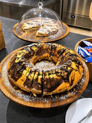 Ein Gugelhupf auf einer Holzbrett, ein abgedeckter Kuchen auf einem Präsentierteller und eine Mikrowelle im Hintergrund.