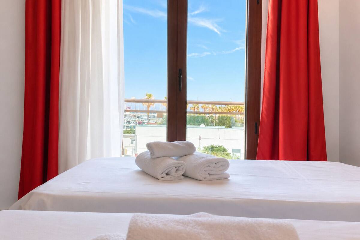 Une chambre d’hôtel avec un lit, du linge de lit blanc et une vue sur un port de plaisance, visible à travers une grande fenêtre ornée de rideaux rouges.