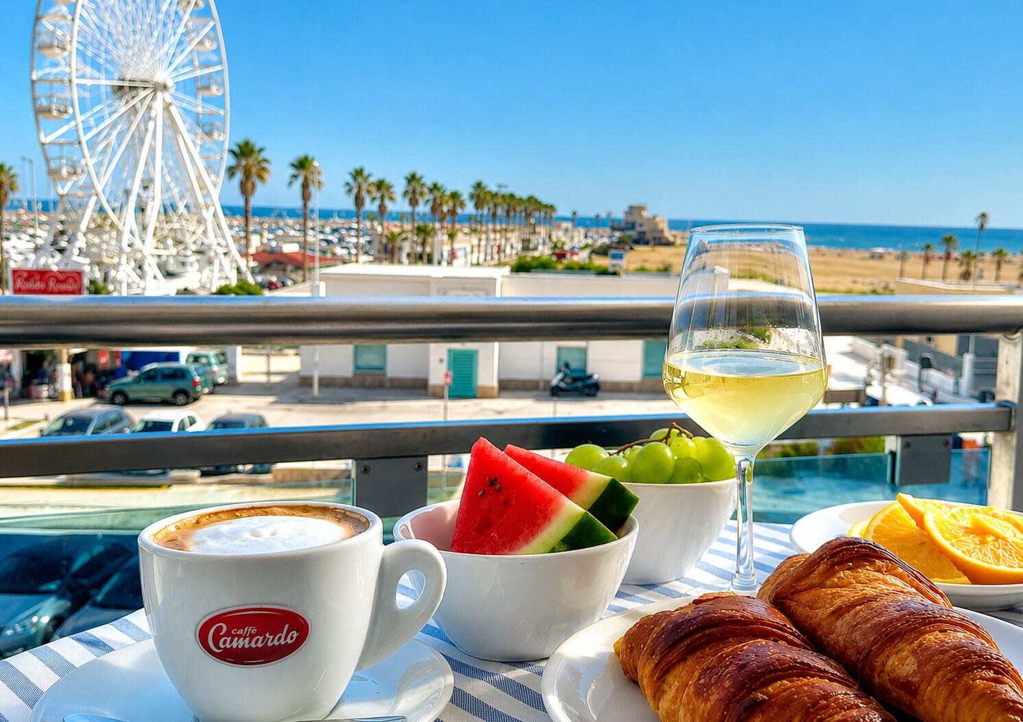 A balcony with a view of a beach, a Ferris wheel, and a table with a croissant, fruit, and a glass of white wine.