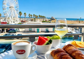 A balcony with a view of a beach, a Ferris wheel, and a table with a croissant, fruit, and a glass of white wine. - B&B MARINA SLEEP AND SHIP (Rodi Garganico)