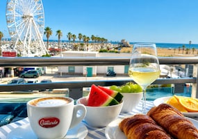 Un balcón con vista a una playa, una noria y una mesa con un croissant, fruta y una copa de vino blanco.