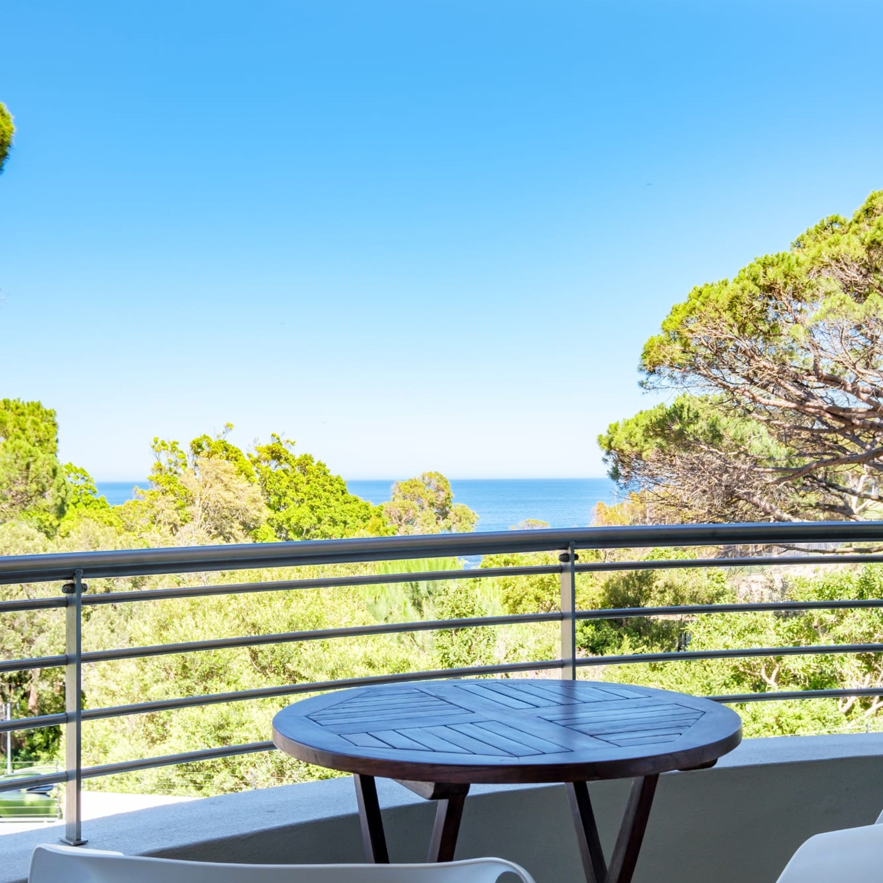 A balcony with a round table and chairs, overlooking a coastal landscape with trees and the sea.
