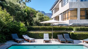 A swimming pool with lounge chairs and a sun umbrella, surrounded by greenery and a building with large windows. - Delightful Rental w Sea Views Vadi Apartment (Cape Town)