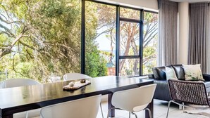 A modern living room with a dining area, large windows, and a view of trees. - Delightful Rental w Sea Views Vadi Apartment (Cape Town)