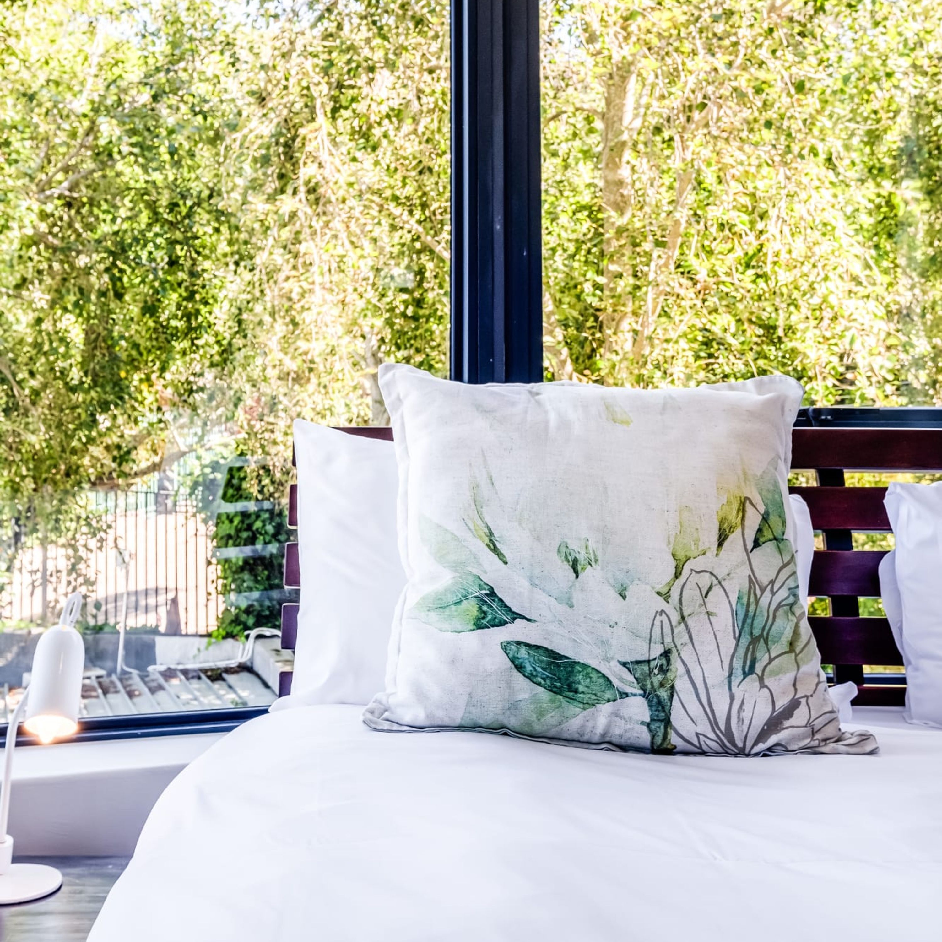 A bed with a white pillow featuring a leaf pattern, a wooden headboard, and a view of green foliage through a window.