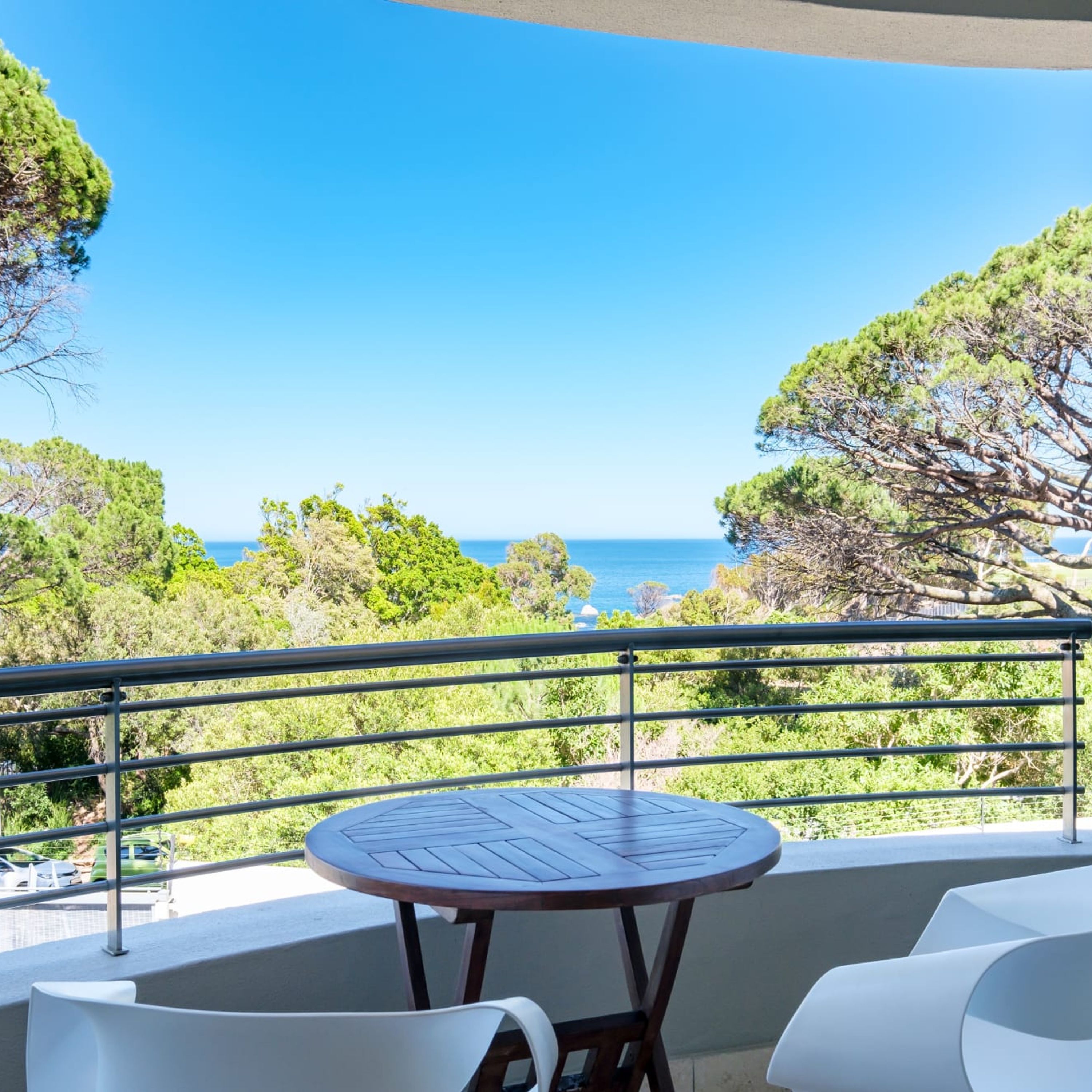 A balcony with a round table and two white chairs, overlooking a coastal landscape with trees and the sea.