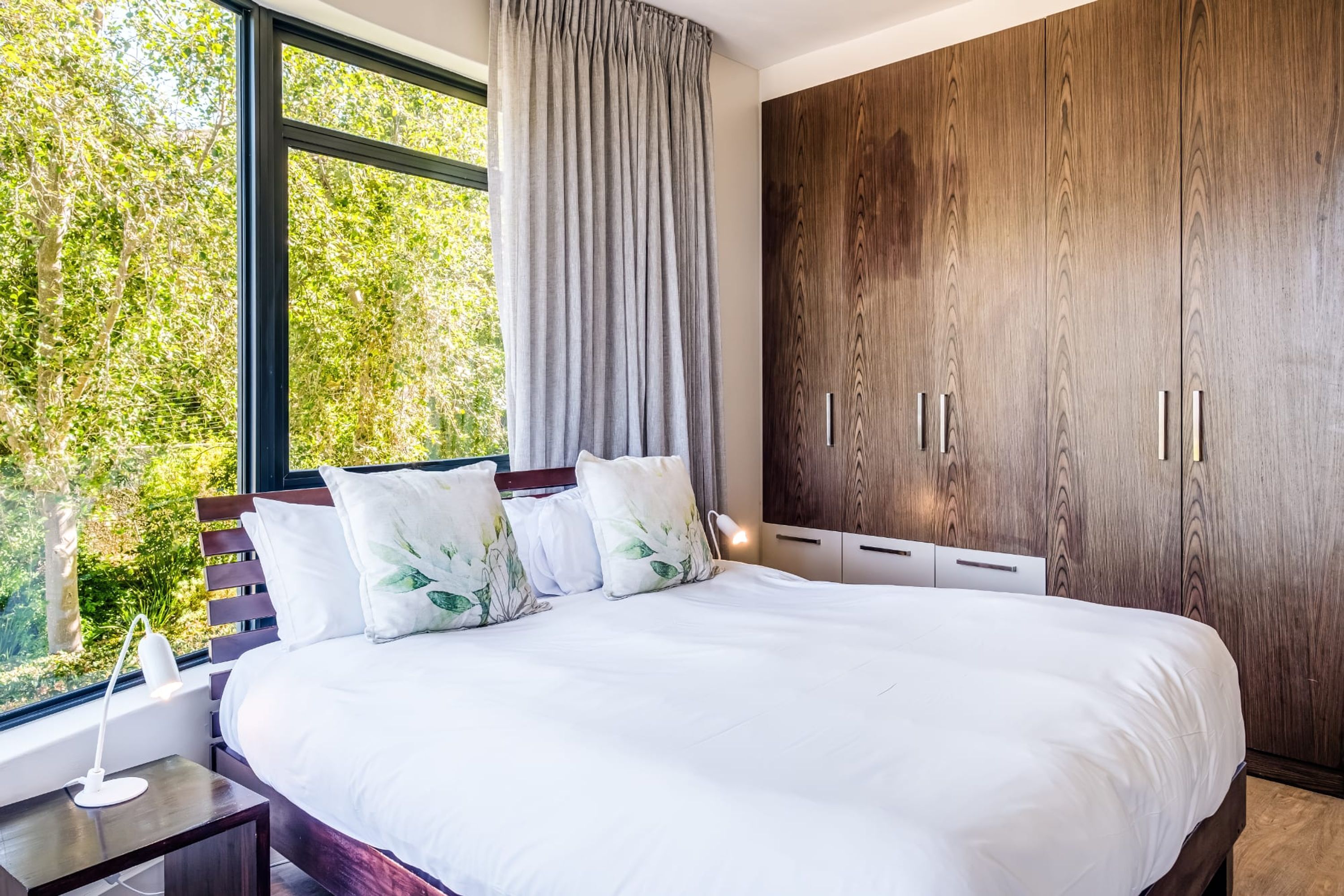 A bedroom with a large bed, wooden wardrobe, and a window with a view of greenery.