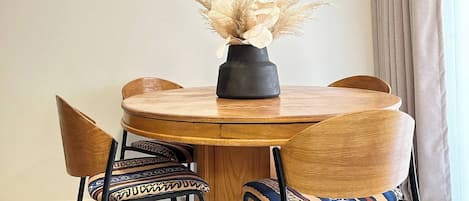 A round wooden dining table with a black vase of dried flowers, surrounded by four chairs with patterned cushions.