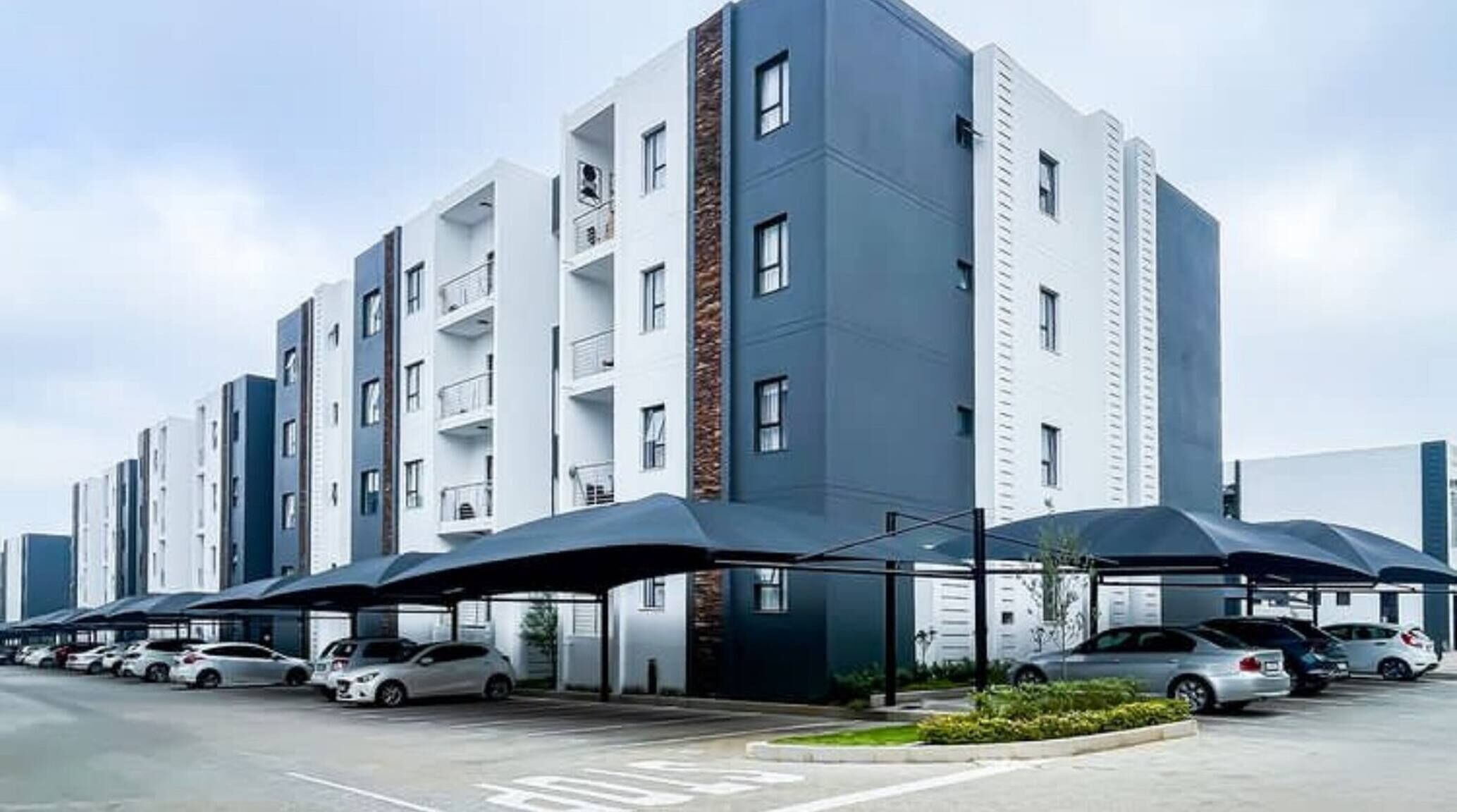 A multi-story residential building with a modern design, featuring balconies and a parking area with cars.