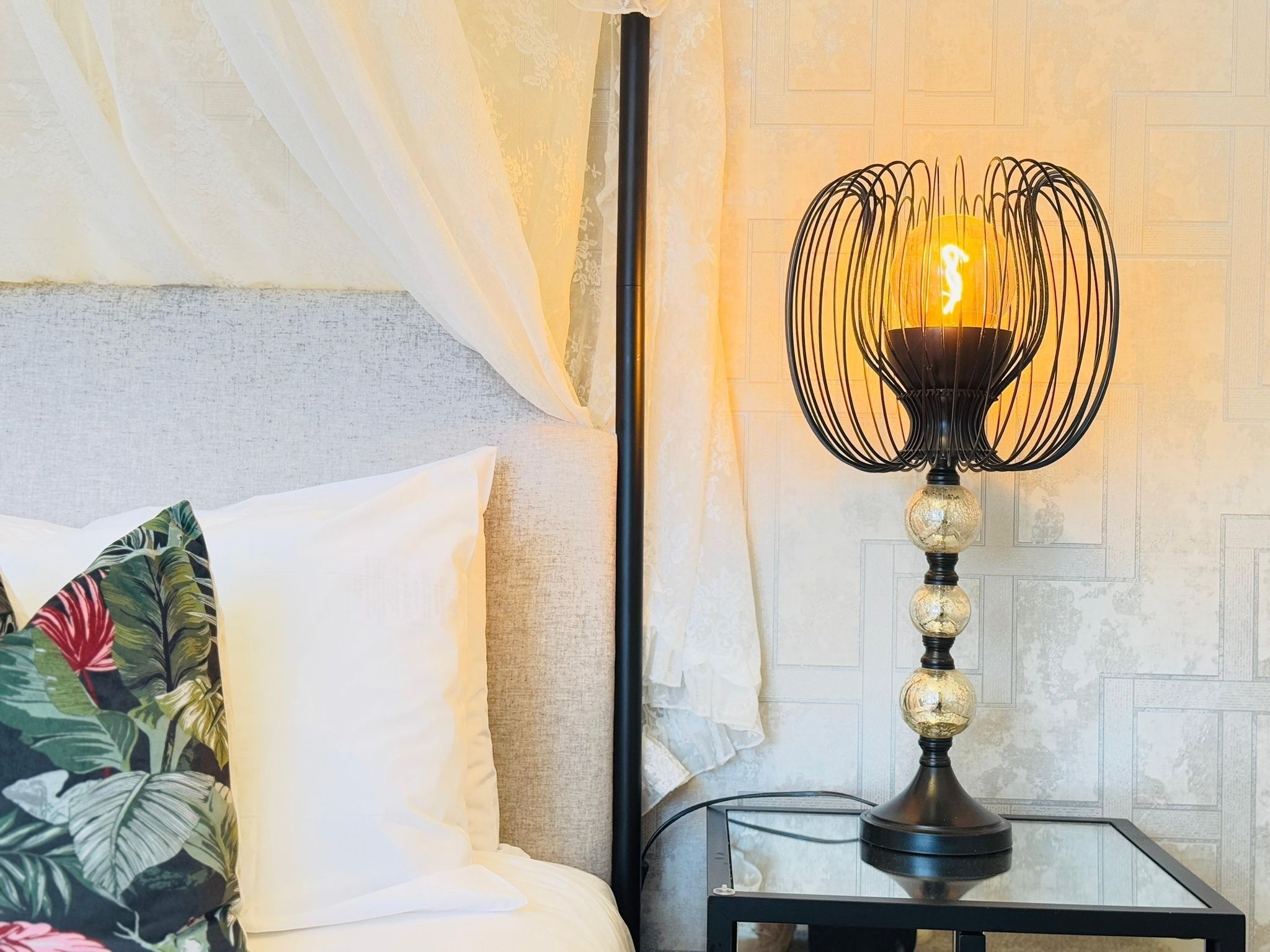 A bed with white linens and a floral pillow, a bedside table with a lamp, and a wall with a geometric pattern.