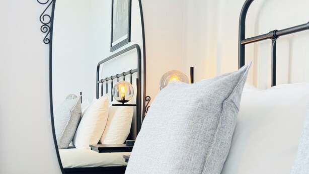 A neatly made bed with white and grey bedding, a bedside table with a lamp, and a mirror reflecting the room. - Skyline View Apartment (Edinburgh)