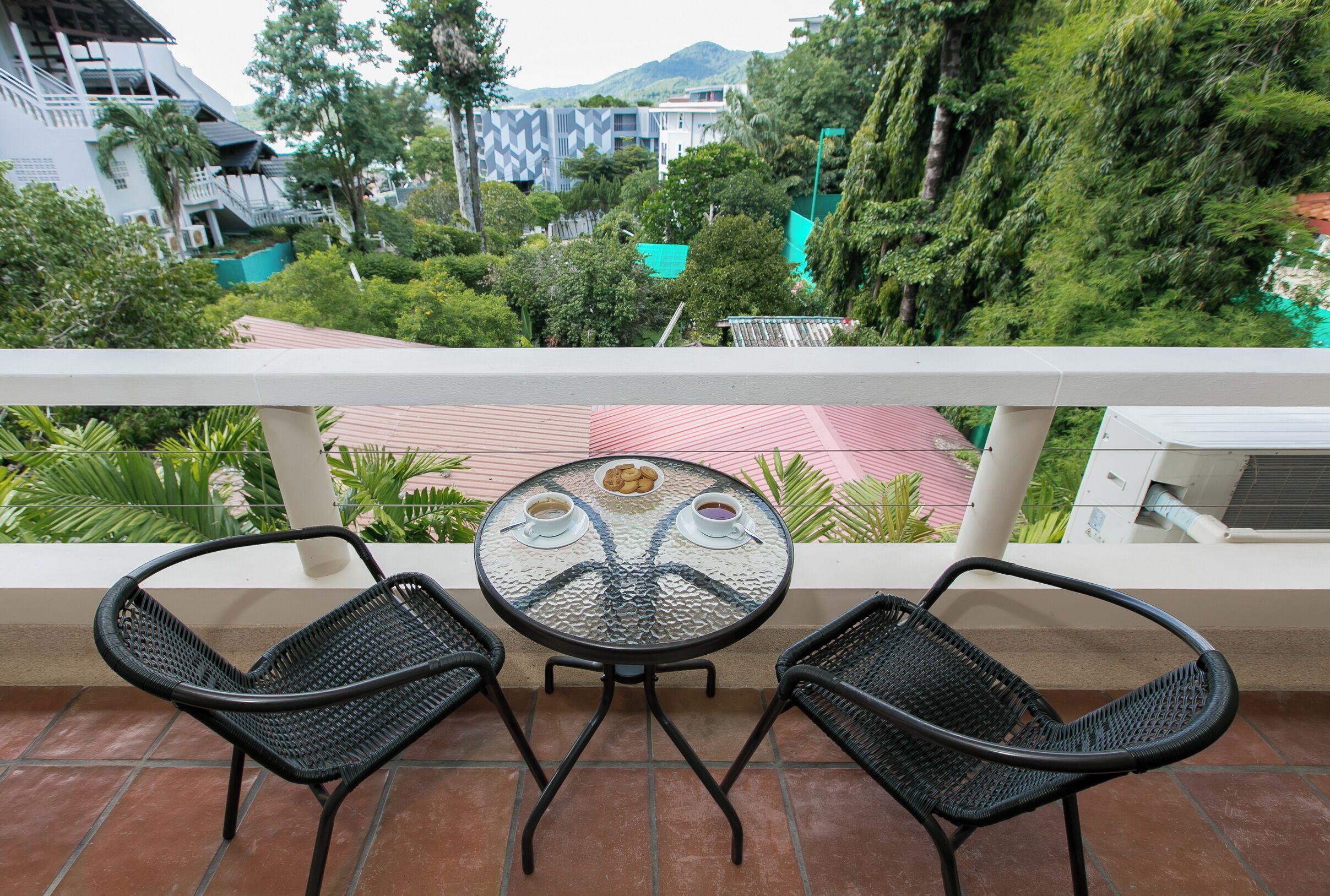 Un balcón con dos sillones de mimbre negro y una mesa de vidrio con un centro decorativo, con vistas a un jardín exuberante y edificios.