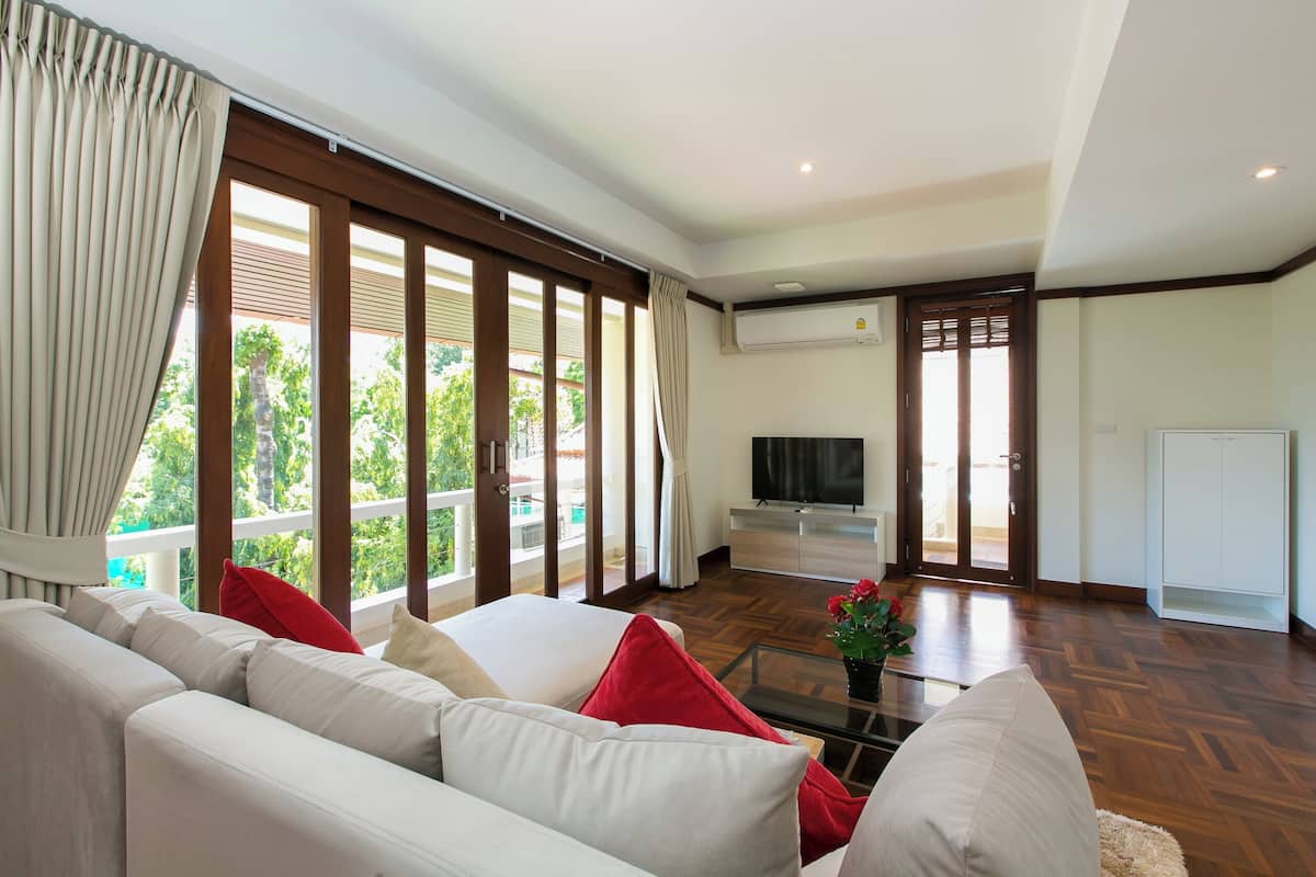 A spacious living room with a large sofa, a television, and a view of the outdoors through large windows.