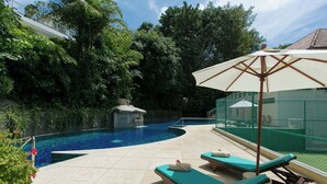 Piscina con sillones de descanso y una sombrilla, rodeada de vegetación y una zona delimitada por una cerca.