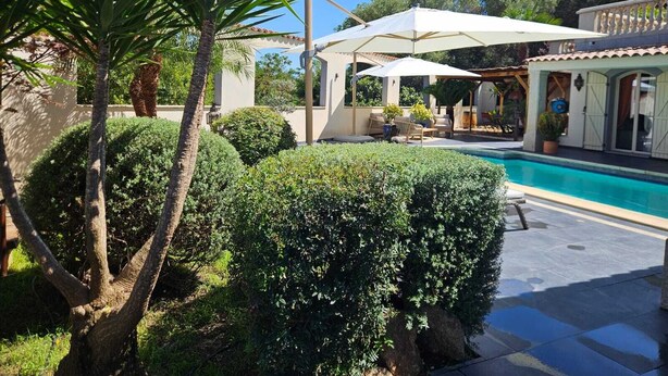 A pool area with a covered seating area, a pool, and surrounding greenery.