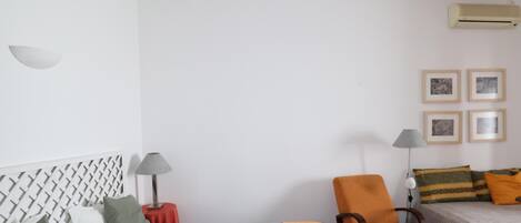 A simple, clean room with a bed, a chair, a small table, and a wall-mounted air conditioner.