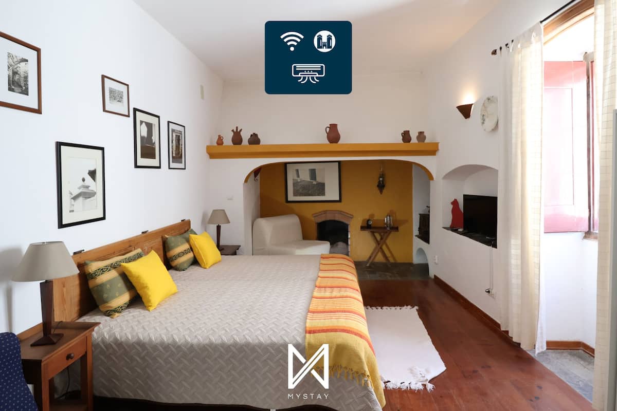 A bedroom with a bed, bedside tables, a lamp, a fireplace, and a shelf with decorative items.