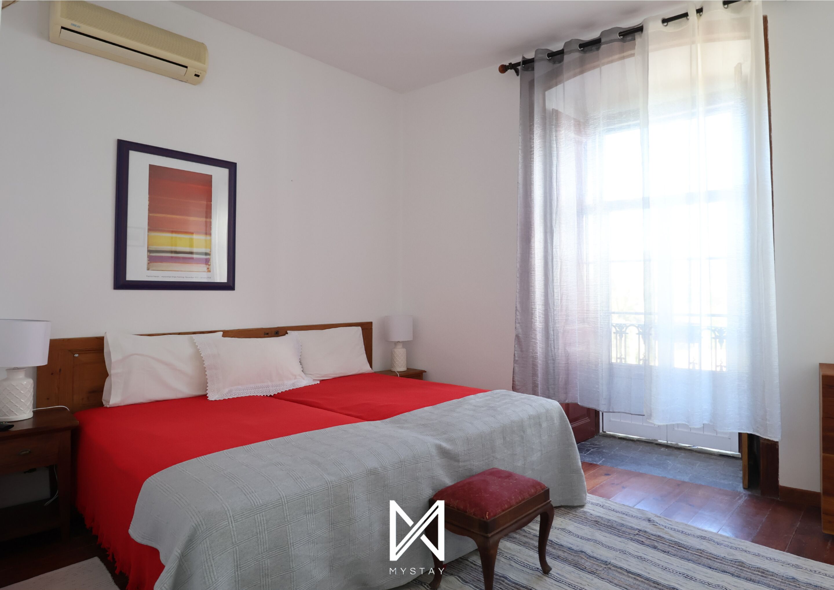 A hotel room with a bed, a bedside table, a lamp, a window with sheer curtains, and a framed picture on the wall.