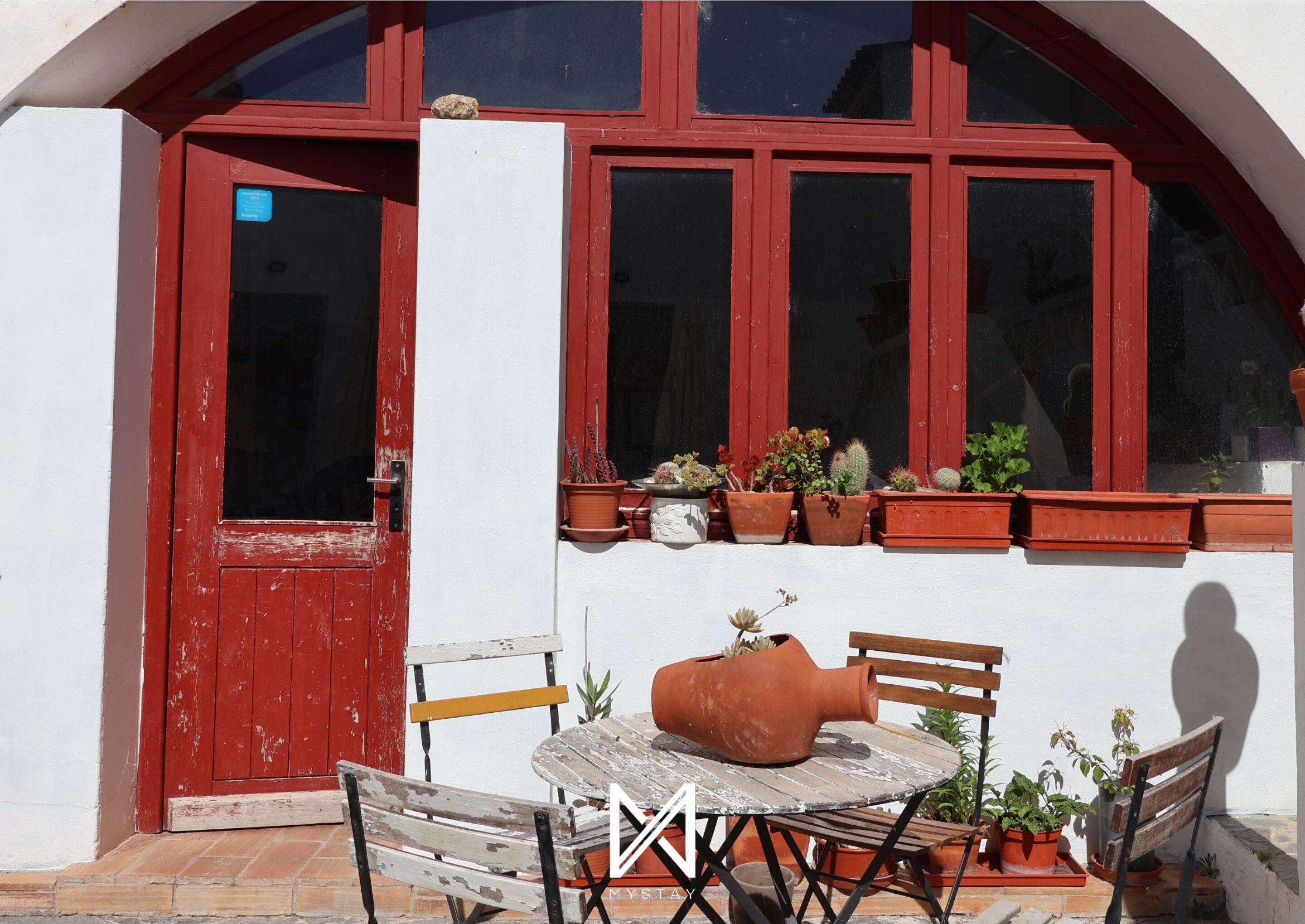 Une porte rouge avec une fenêtre au-dessus, une petite table avec des chaises et des plantes en pot sur le rebord de la fenêtre.