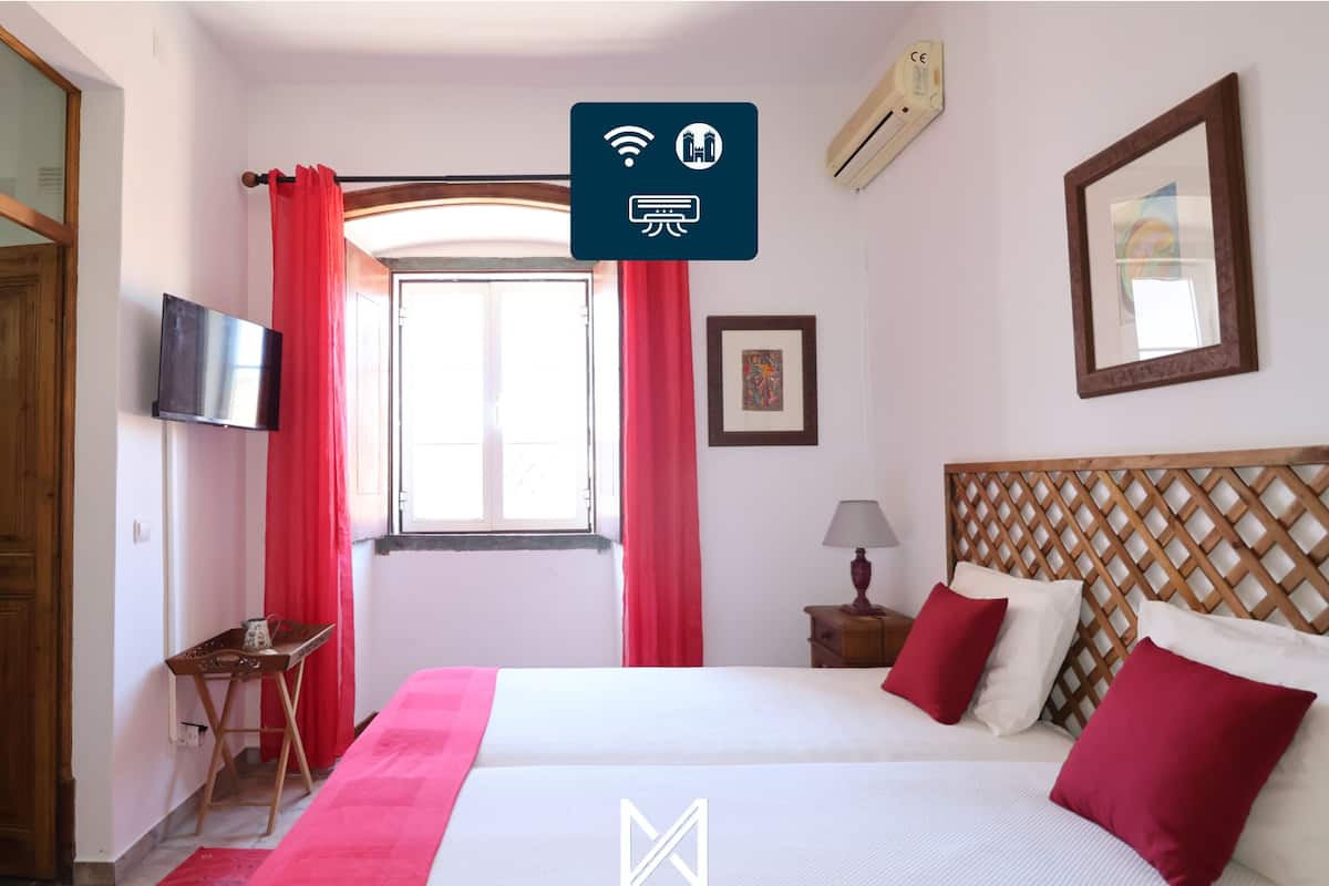 A hotel room with a bed, a TV, a window with red curtains, a wooden headboard, and a framed picture on the wall.