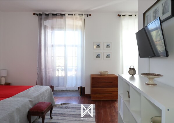 Une chambre à coucher comprenant un lit, une commode, un téléviseur et une fenêtre avec des rideaux légers.