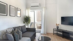 Ein modernes Wohnzimmer mit einem grauen Sofa, einem runden Couchtisch, einem Fernseher auf einem schwarzen Standfuß und einem Balkon mit weißen Vorhängen.