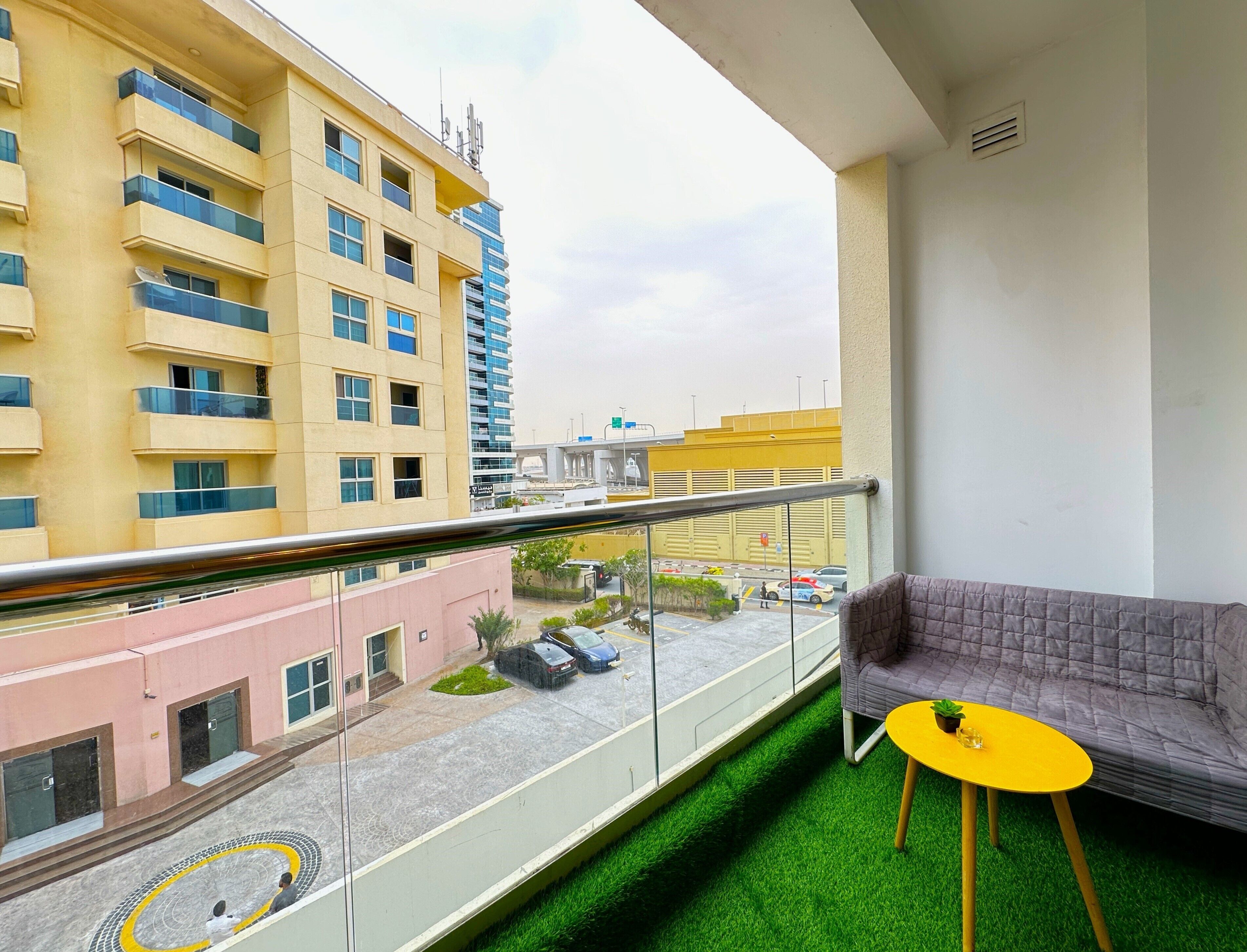 A balcony with a grey sofa, a yellow table, and a view of a building with multiple floors and balconies.