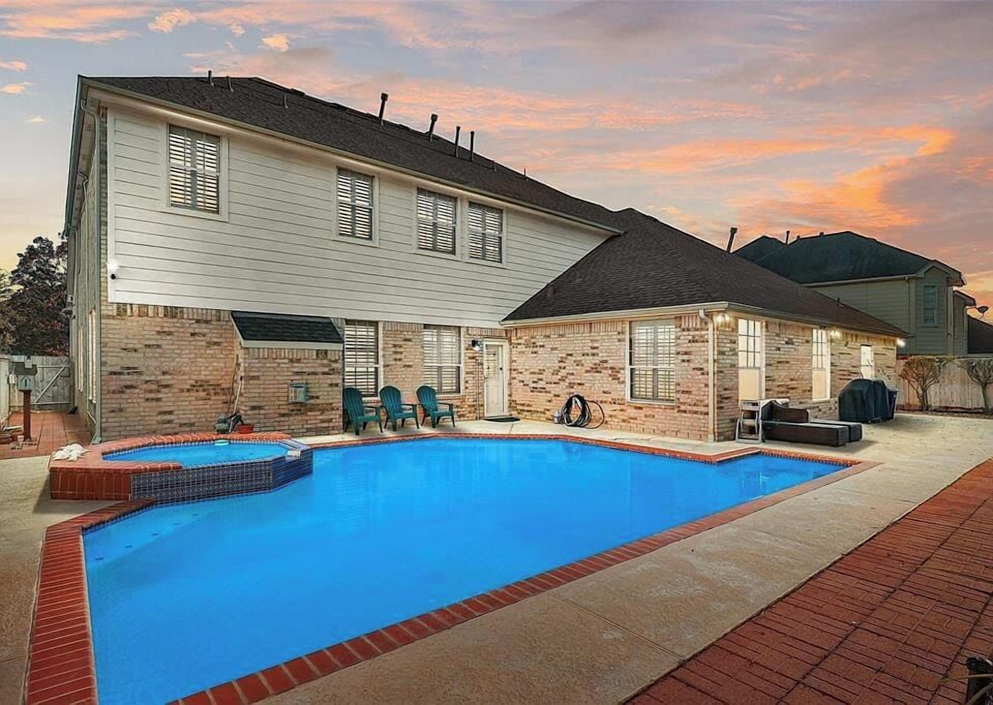 A two-story house with a swimming pool, brick exterior, and a covered patio area with chairs.