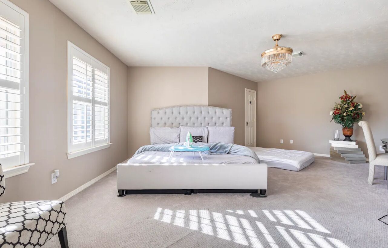 Une chambre avec un grand lit, un lustre, une fenêtre avec des stores et une plante décorative.