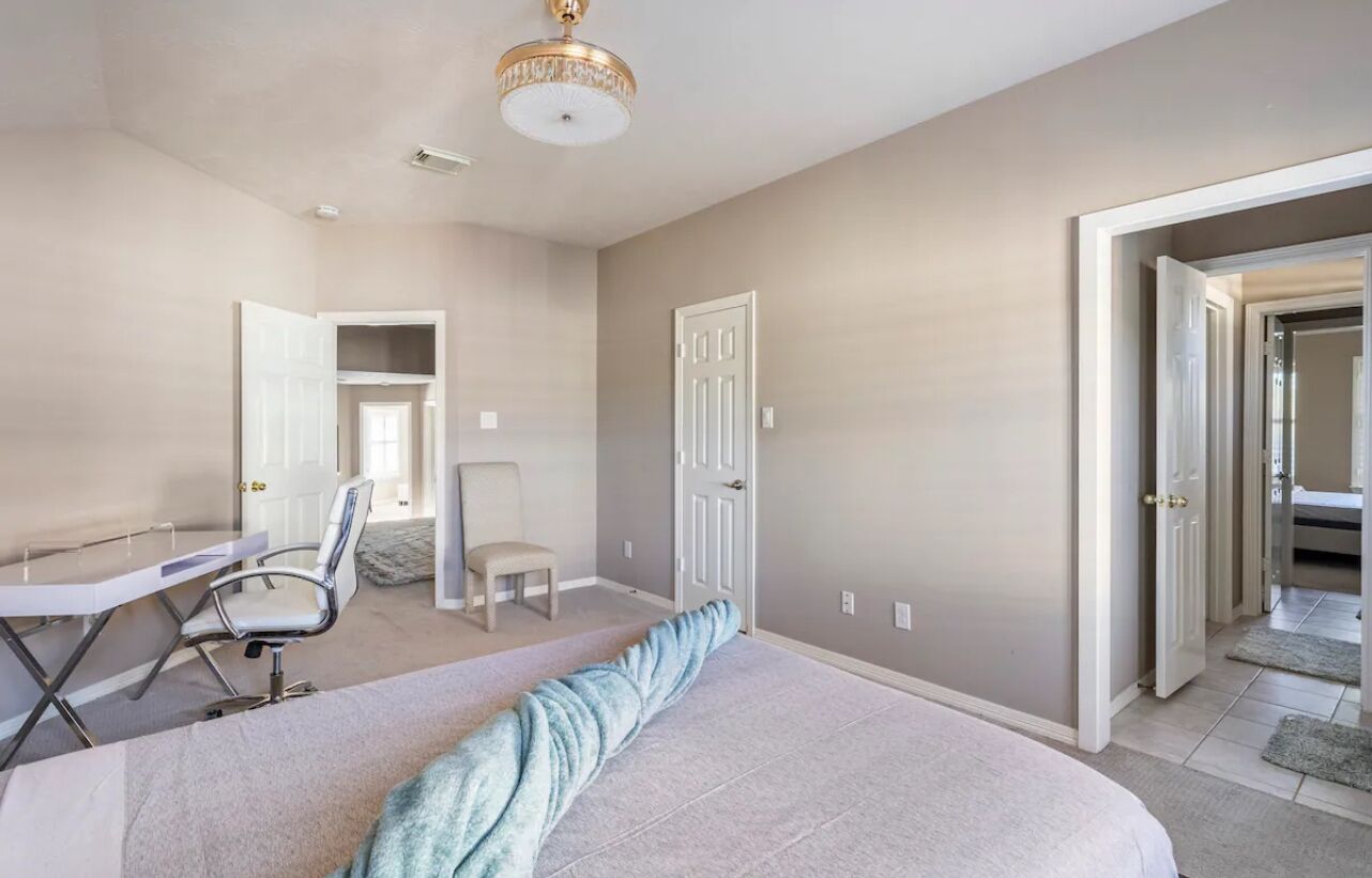 A bedroom with a large bed, a desk, and two chairs. There are two doors and a ceiling light.