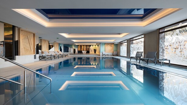 A modern indoor swimming pool with a clear blue water surface, sunlit ceiling, and lounge chairs along the poolside. - MGM Hôtels & Résidences – Résidence Akoya (Les Avanchers-Valmorel)