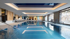Une piscine intérieure moderne, avec une eau d’un bleu cristallin, un plafond baigné de lumière et des chaises longues disposées le long du bord de la piscine.
