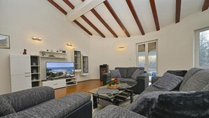 Een moderne woonkamer met grijze banken, een flatscreen televisie, houten balken in het plafond en een groot raam.