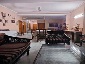 A living room with a sofa, a coffee table, a dining table, and chairs. There is a ceiling fan, a clock, and framed pictures on the walls. - Room-home Stay Bed And Break Fast (New Delhi)