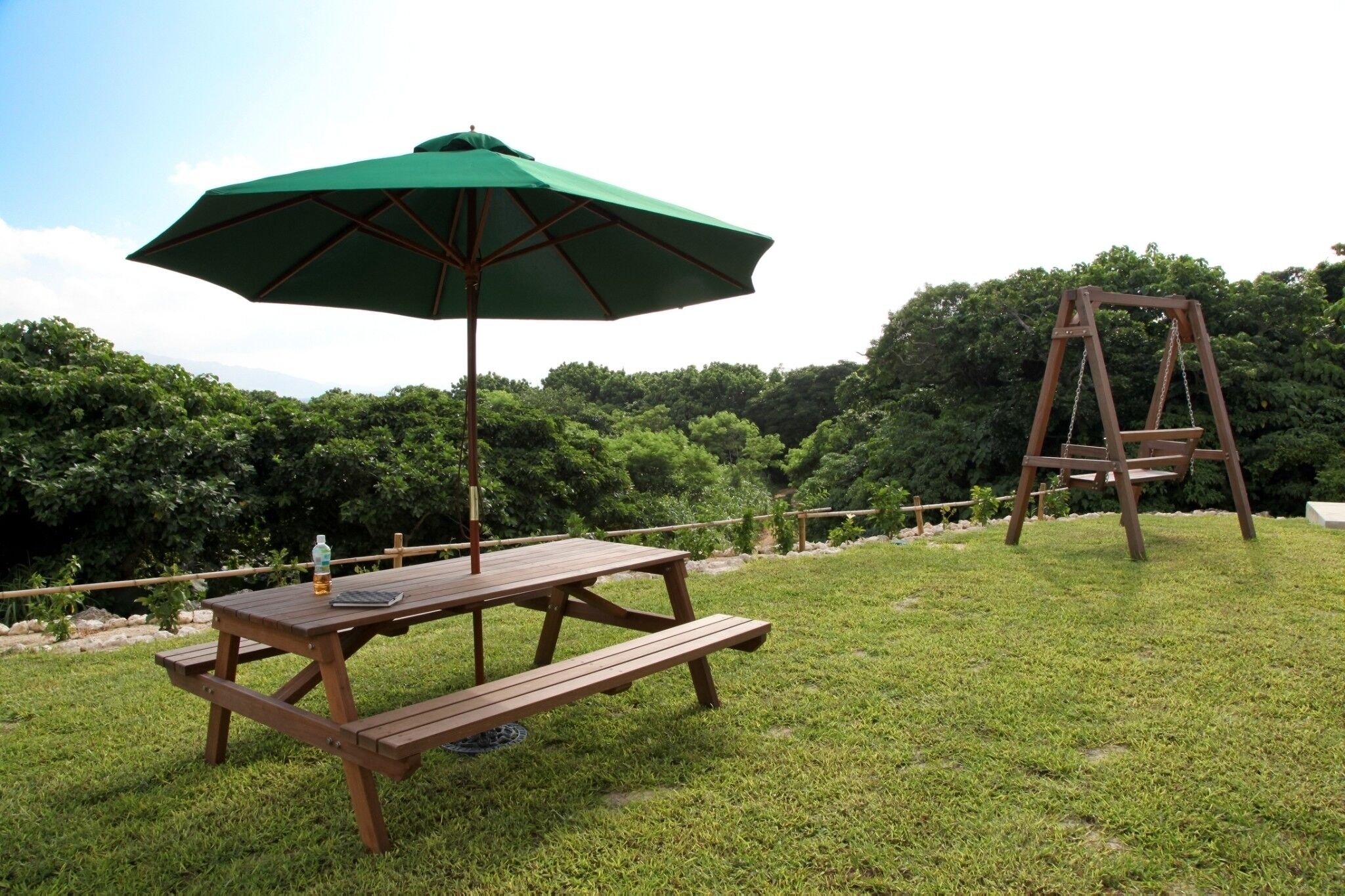 A picnic table with a green umbrella and a wooden swing set in a grassy area with trees in the background.