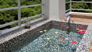 A rooftop infinity pool with mosaic tiles and floating flowers, surrounded by white railings and a view of lush greenery and distant mountains.