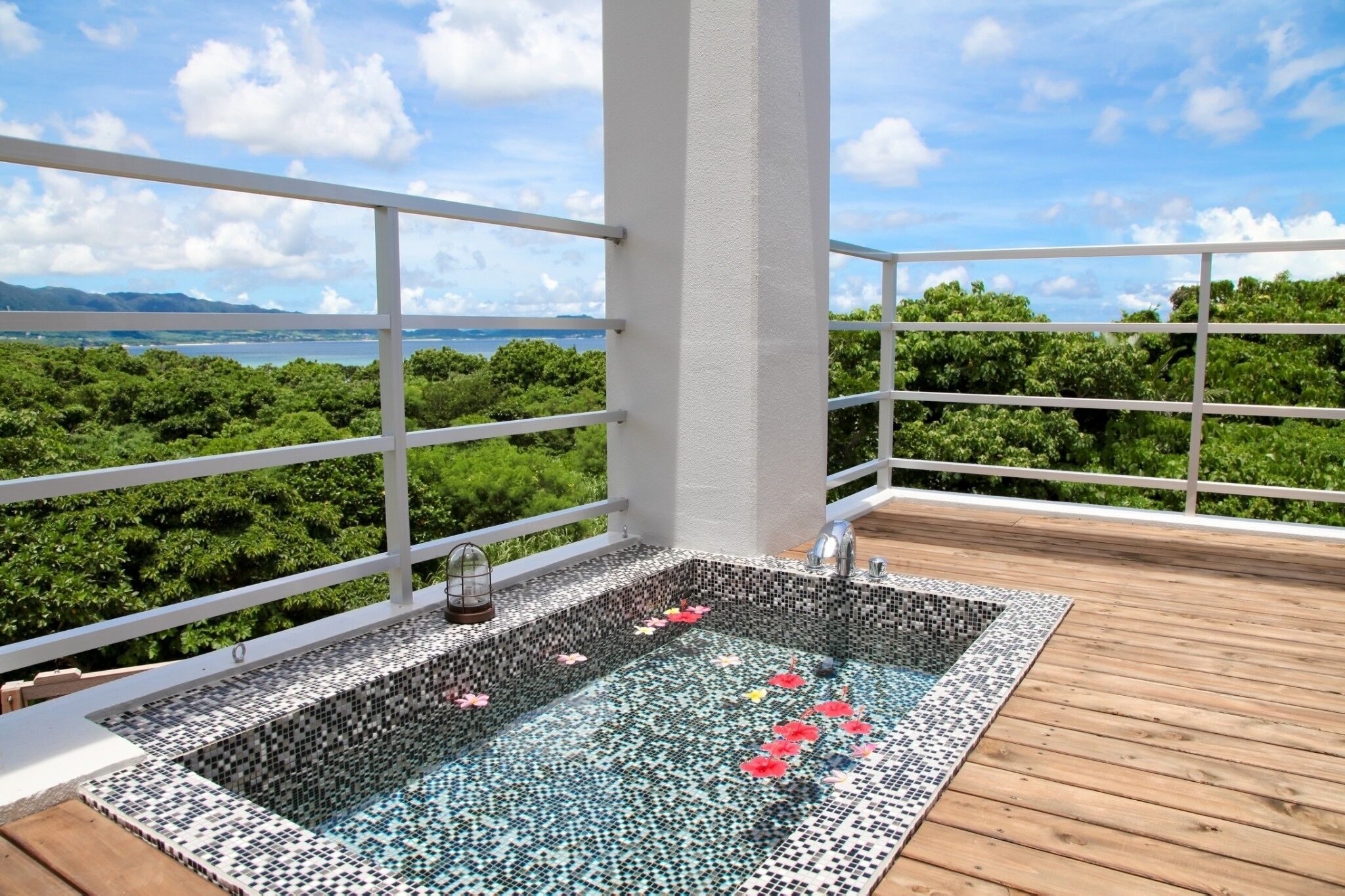 Ein Pool auf dem Dach mit Mosaikfliesen und einer Holzdachterrasse, umgeben von weißem Geländer und üppiger Vegetation.