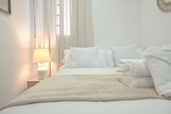 Un lit bien fait, avec du linge de lit blanc et un plaid beige, une lampe de chevet et un objet décoratif fixé au mur.