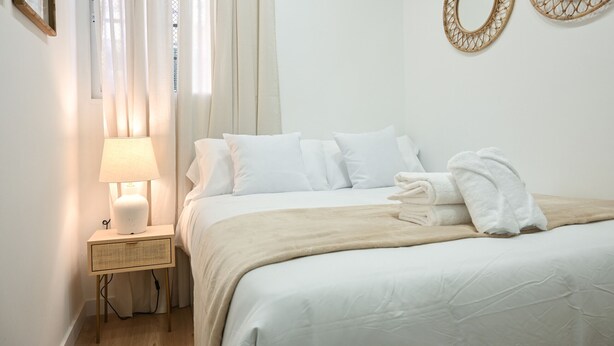 A neatly made bed with white linens and a beige throw blanket, a bedside lamp, and two decorative mirrors on the wall. - Charming one-bedroom apartment in Madrid, close to the Tetuán metro (Madrid)