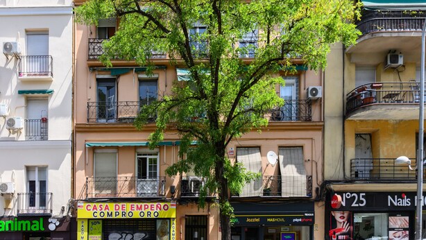Un edificio de varios pisos con balcones, un árbol en primer plano y locales comerciales, entre ellos una sala de manicura y una tienda con un letrero amarillo.