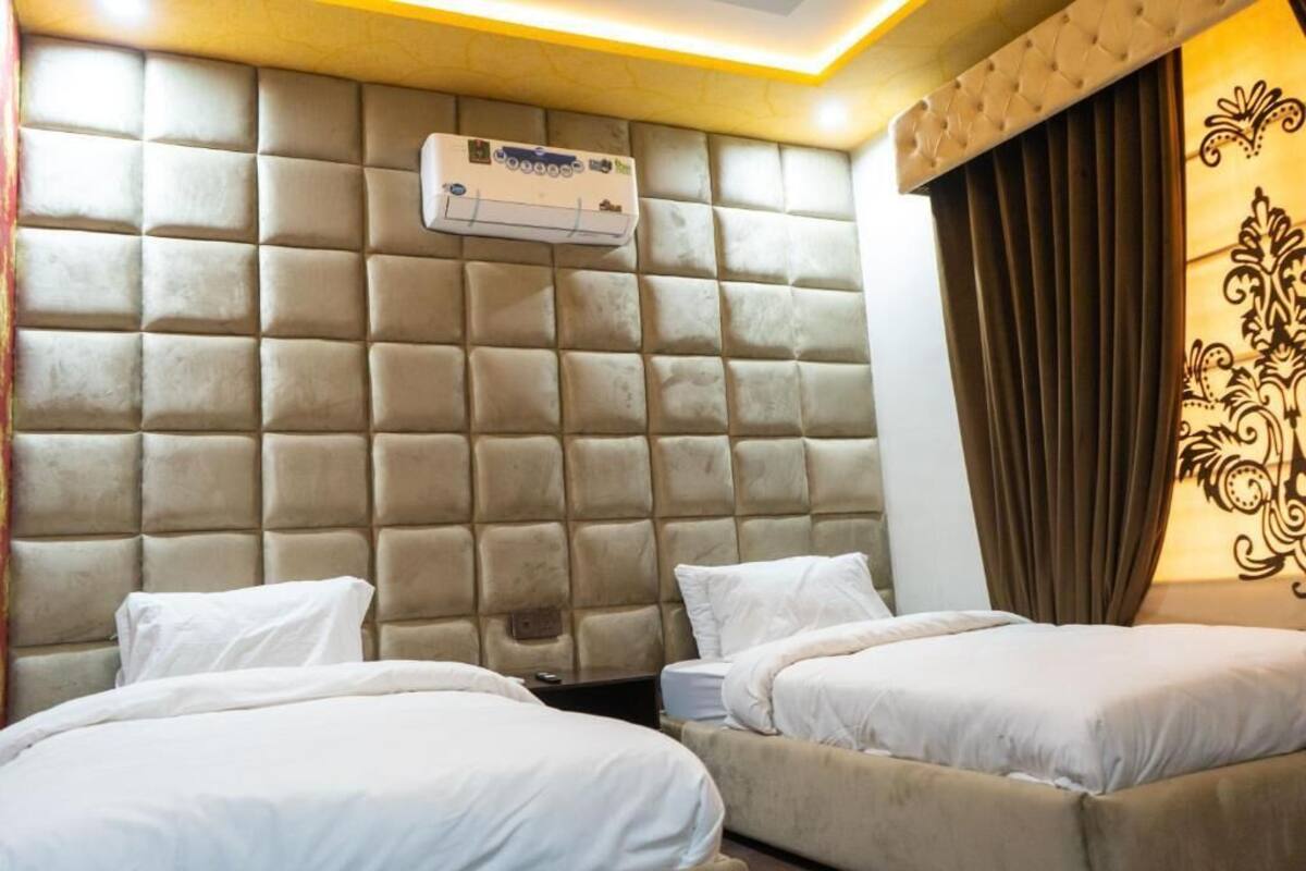 A hotel room with a tufted headboard, two beds with white bedding, an air conditioning unit, and a decorative wall panel.
