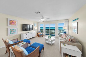 A spacious living room with a large TV, comfortable seating, a small table, and a balcony with a view of the ocean.