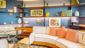 Un salon moderne avec un canapé blanc aux lignes courbes, des coussins à motifs orange et une table basse en verre. On y trouve des étagères remplies de livres, d’objets décoratifs et de tableaux encadrés.