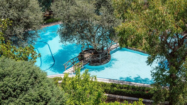 Une vue aérienne d’une piscine entourée d’une végétation luxuriante, avec une fontaine centrale et une terrasse en bois.