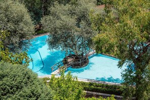 An aerial view of a swimming pool surrounded by dense greenery, with a central water feature and a wooden deck.