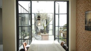 Un comedor con una mesa grande y sillas, una lámpara colgante, y vistas a un espacio exterior con plantas y un muro de ladrillo.