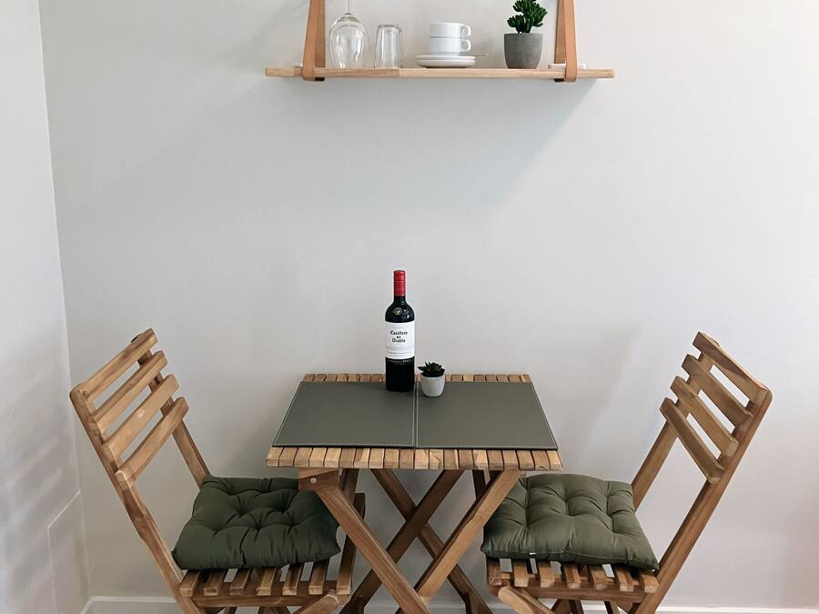 Un pequeño comedor con una mesa cuadrada, dos sillas de madera, una botella de vino y una pequeña planta en maceta sobre un estante.