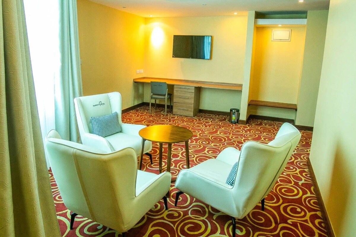 A hotel room with a desk, two chairs, a small table, a TV on the wall, and a patterned carpet.
