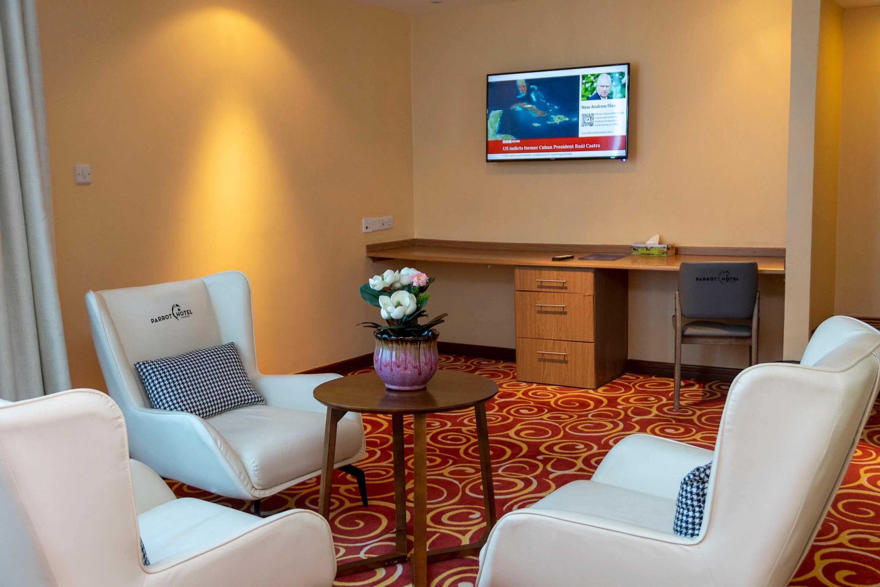 Habitación de hotel con dos sillones de cuero blanco, una mesita con un florero, un escritorio con una computadora y un televisor en la pared mostrando noticias.
