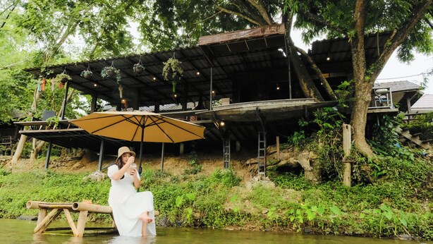 A woman in a white dress sitting on a wooden bench by a river, under a large umbrella. - Pastela Doi Saket Villa & Spa (Doi Saket)